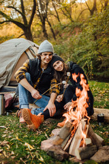 Young loving couple of tourists relaxing near the fire in the nature..Handsome man and beautiful woman are roasting marshmallows over the fire near tent in camping. Instagram stories picture.