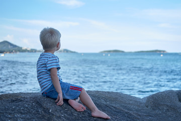 Portrait of little handsome boy sitting on rock near sea water, looking dreamily into the distance. Thailand