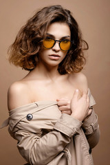 beauty fashion model with clean skin and curly hair in biege cloak stretch on biege background, model in fashion glasses, bare shoulders