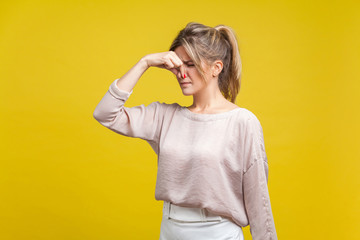 Unpleasant smell. Portrait of young dissatisfied woman with fair hair in casual beige blouse standing with closed eyes, pinching her nose with disgust. indoor studio shot isolated on yellow background