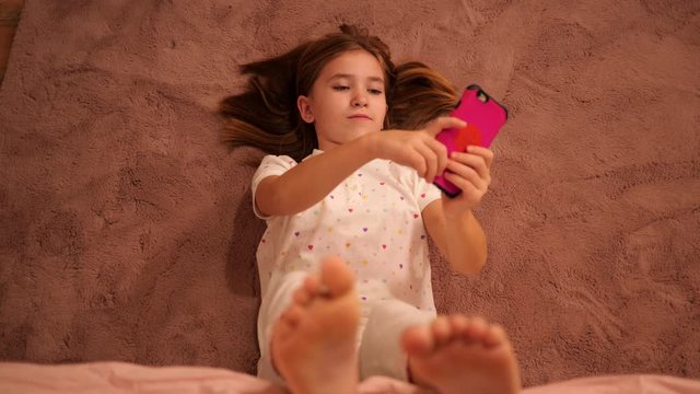 Kid girl of school age, lie on a floor in bedroom texting messages to friends using smartphone having fun before go to bed