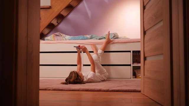 Kid girl of school age, lie on a floor in bedroom texting messages to friends using smartphone having fun before go to bed