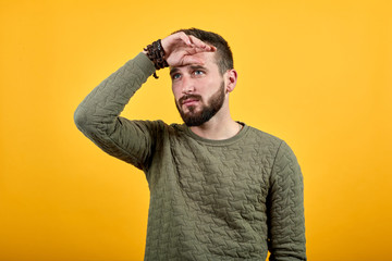 Handsome serious caucasian man over isolated orange background keeping hands on forehead, looking into distance wearing casual green clothes