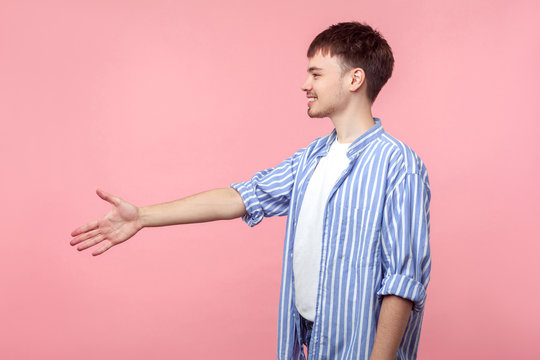 Side View Of Positive Friendly Brown-haired Man With Small Beard And Mustache In Casual Striped Shirt Holding Out His Hand To Side, Giving Handshake. Indoor Studio Shot Isolated On Pink Background