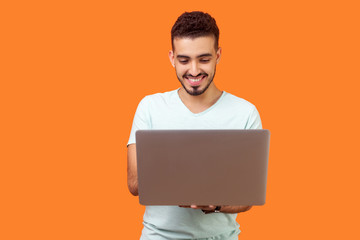 Portrait of cheerful young brunette man with beard in white t-shirt typing email on laptop and...