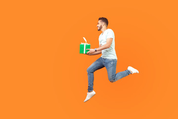 Full length portrait of excited happy brunette man in sneakers and denim outfit flying or running in air with gift box, holiday present, hurrying for sale. studio shot isolated on orange background