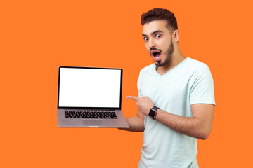 Portrait of excited shocked surprised brunette man with beard in white t-shirt pointing at laptop with blank screen, empty place for internet advertising. studio shot isolated on orange background - obrazy, fototapety, plakaty