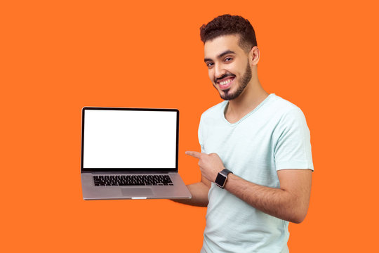 Portrait of positive joyful brunette man with beard in white t-shirt standing holding laptop with blank screen and smiling at camera, internet advertising. studio shot isolated on orange background