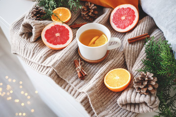 Fototapeta na wymiar Christmas cozy winter home decor. New year still life: cup of citrus tea, oranges, grapefruits, cinnamon sticks, pine cones, fir branches, knitted sweater, glowing led garland lights.
