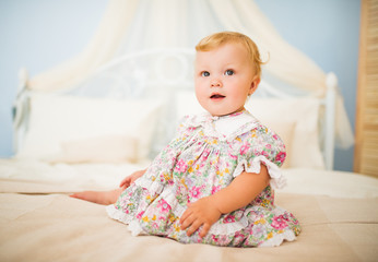 Portrait of a cheerful pretty fair-haired chubby little girl in a dress sitting on a large bed. Healthy and happy children concept. Concept for advertising baby products