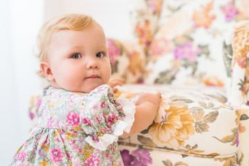 Portrait of a charming little girl in a beautiful dress standing near a chair with a floral print. Concept of the first steps and cognition of the world of young children. Advertising concept