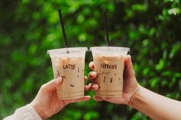 Lover couple hands holding and showing a glass cup of ice coffee latte and ice espresso outdoors on green leaf background.Vintage film tone.