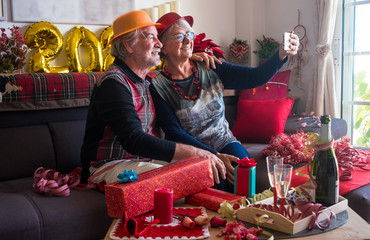 Two happy senior people with  party hat sitting on the sofa and looking at mobile phone for a selfie. Toasting with a sparkling wine. One elderly couple