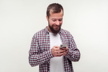 Mobile communication. Portrait of cheerful bearded man in casual plaid shirt using cell phone chatting in social network and smiling, reading good news. indoor studio shot isolated on white background