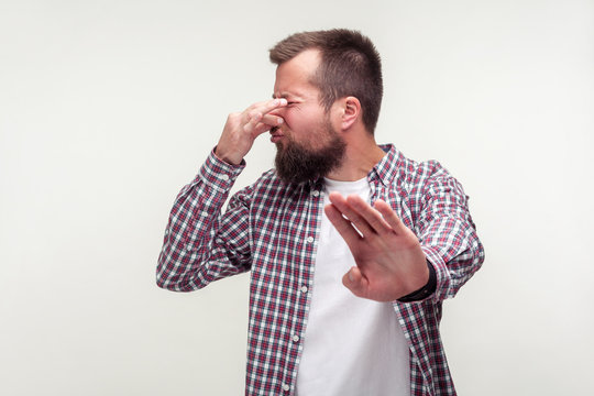 Portrait of confused bearded man in casual plaid shirt showing stop gesture and pinching nose with disgust grimace, feeling repulsion to unpleasant smell. studio shot isolated on white background