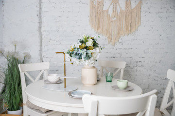 Stylish kitchen in white, pastel colors. Style minimalism. Vase with flowers, white table, plants,glasses, plates,dishes. Trendy interior with white furniture, table, brick walls. loft apartment