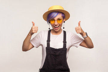 Like, well done! Portrait of stylish wonderful hipster girl with violet short hair in sunglasses and hat showing thumbs up gesture, success sign and smiling. isolated on white background, studio shot