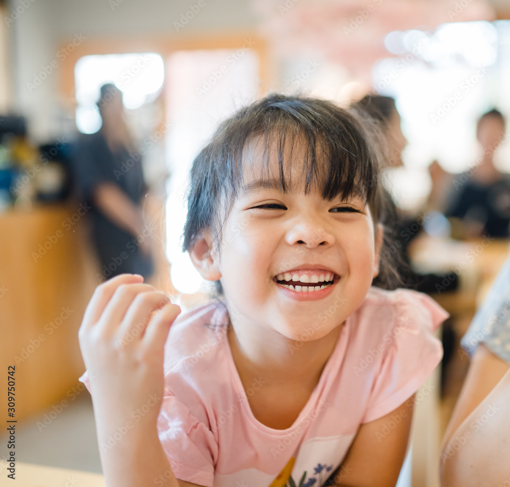 Wall mural Happy Little asian girl child showing front teeth with big smile and laughing in restaurant, she talk with mom: Healthy happy funny smiling face young adorable lovely female kid.