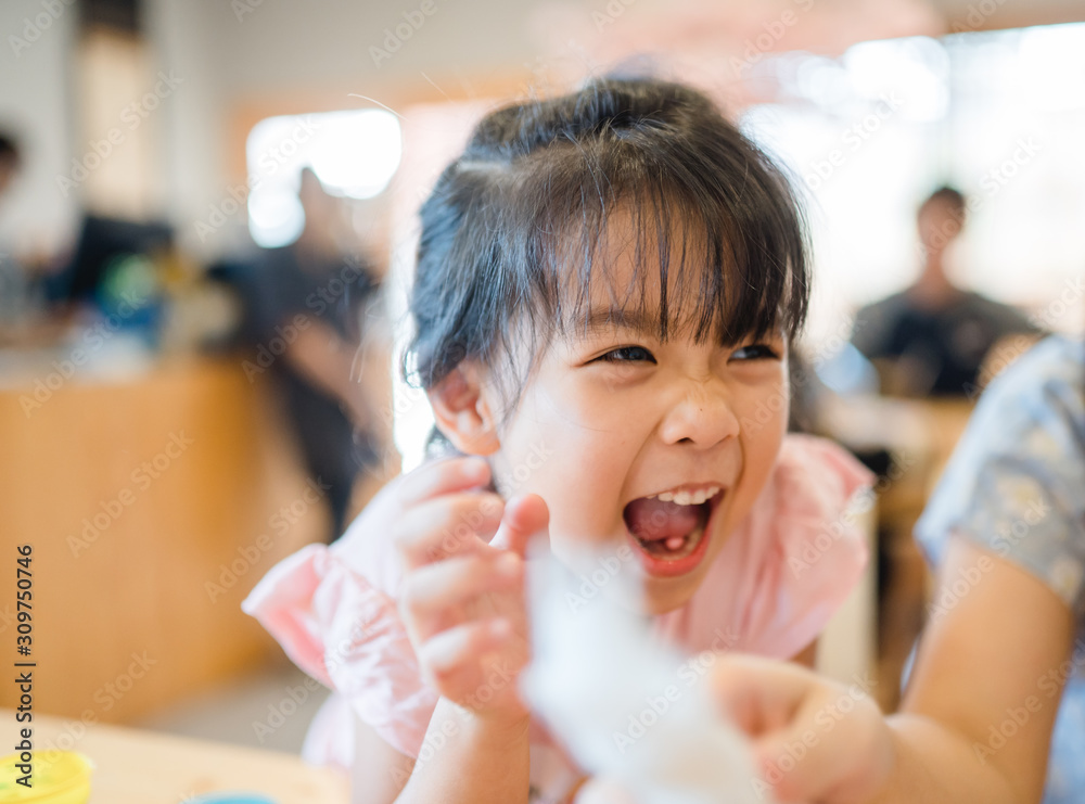 Wall mural Happy Little asian girl child showing front teeth with big smile and laughing in restaurant, she talk with mom: Healthy happy funny smiling face young adorable lovely female kid.