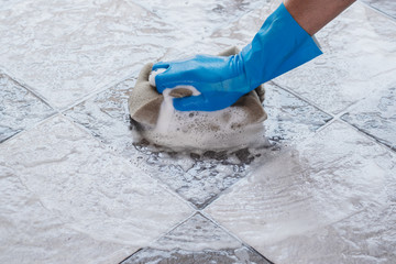 Hand of man wearing blue rubber gloves is using a sponge cleaning the tile floor.