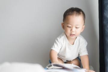 Naklejka premium 2 years little asian toddler boy in white t-shirt reading bible picture story for kid in the morning.Hands holding on a Holy Bible in bed room concept for Education bible study, faith and religion.