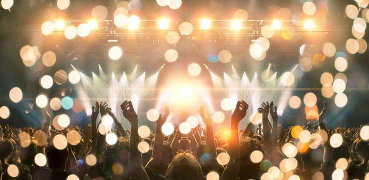 Photo of a concert hall with people silhouettes clapping in front of a big stage lit by spotlights. Shot is taken from concert crowd point of view, lens flare is visible.