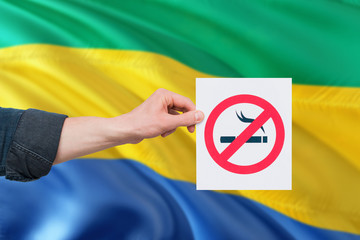 Gabon health concept. Hand holding paper with no smoking sign over national waving flag. Quit smoke...