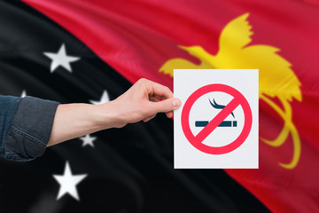 Papua New Guinea health concept. Hand holding paper with no smoking sign over national waving flag. Quit smoke theme.