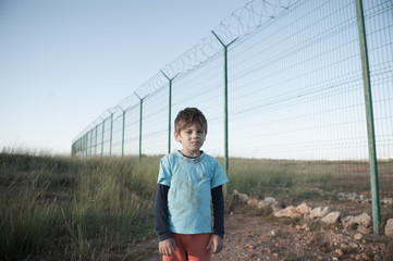 poor little caucasian refugee boy in dirty clothes standing near high fence with barbed razor wire...