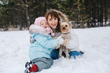 Obraz premium On a winter day, a woman and a girl are sitting in the snow against the background of a forest, hugging and holding a Yorkshire Terrier dog in their arms and looking at the camera.