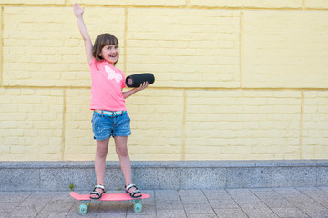 Little happy girl with a skateboard have fun listening to music outdoors.