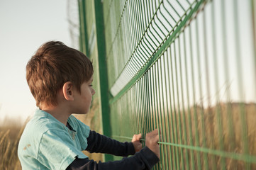 poor dirty little caucasian boy refugee standing near state border fence holding it with hands with...