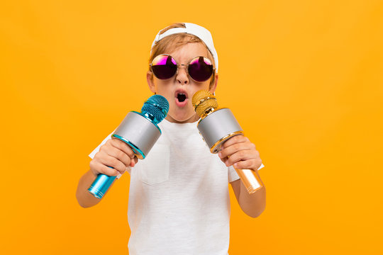 Cheeky blond boy in glasses with two microphones on a yellow background