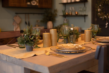 Close-up of beautifully decorated dining table with fir trees in vases and gold candles. Sets of plates for four people.