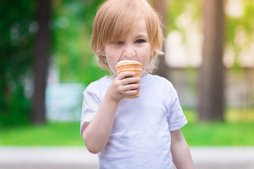 Beautiful baby with blue eyes in white tshirt is eating glass of white ice cream.