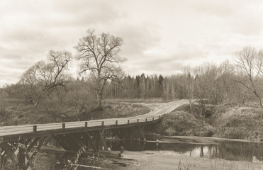 Black and white photograph on a high grain film to produce a retro style. Landscape spring. The riverbank with trees is an old bridge and a country road.