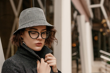 Outdoor close up fashion portrait of young elegant woman wearing trendy checkered bucket hat, stylish cat eye glasses, coat, posing in street of European city. Copy, empty space for text