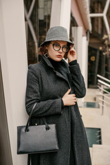 Obraz premium Outdoor fashion portrait of young elegant business woman, model wearing trendy bucket hat, autumn, winter coat, eyeglasses, holding classic black leather bag, handbag, posing in street city