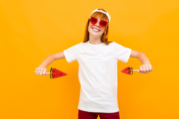 Fototapeta na wymiar smiling european red-haired girl in a white t-shirt and sunglasses with candy in hand on a yellow background.
