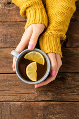 Overhead shot of female hands knitted sweater holding cup of tea with lemon on wooden table