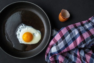 Fried egg in oil in a black cast-iron frying pan. Nearby lies a linen napkin and eggshell on a black background. Close up viev.