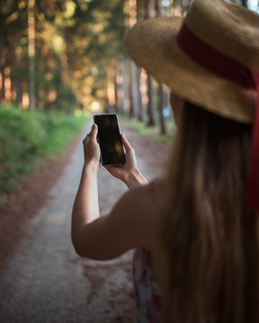 Girl wearing a straw hat taking photo on smart phone in the forest