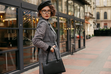 Young elegant business woman, model wearing autumn checkered coat, wrist watch, leather beret, glasses, holding classic bag, handbag, posing in street of European city. Copy, empty space for text