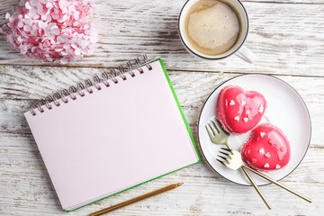 Pink mousse cakes in the shape on a white rustic background. Heart shaped cakes for Valentine's Day or Mother's day. Empty notebook page for text, coffee mug and pink hydrangea. Top view. Flat lay