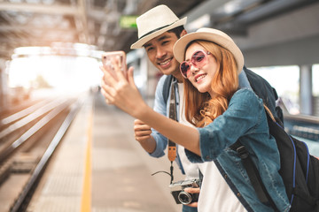 Happy Asian couple traveler holding a mobile phone in station and waiting for train in vacation time.Two Asian Tourists With Backpacks Train travel in Sightseeing City Thailand.