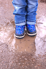 Obraz na płótnie Canvas Kids boots in muddy puddle. Colorful winter outdoor rain shoes on childrens feet in action. A child on a walk stay in the mud. Choosing footwear for an active lifestyle. Autumn rainy weather.