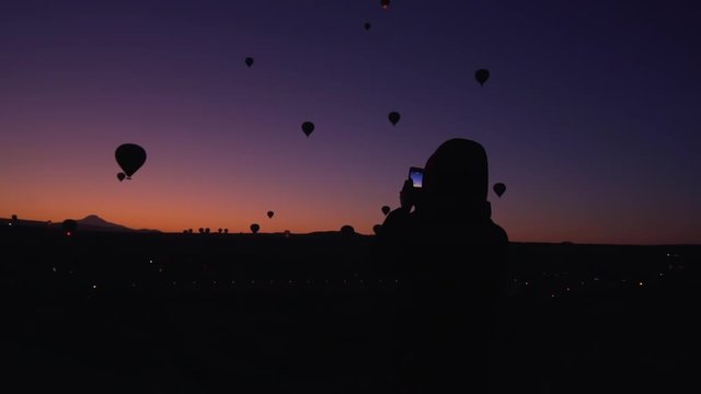 handheld shot of female silhouette taking photos of air balloons at sunrise