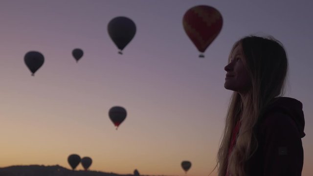 Low key shot of smiley female enjoy looking on beutiful air balloons at sunrise