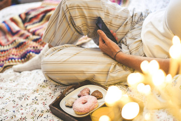 Obraz na płótnie Canvas Young woman lying on bed having breakfast on holidays with cookies and and donut. Morning in a cozy warm bedroom reading Christmas greetings on smartphone. Girl sending message for the new year eve.