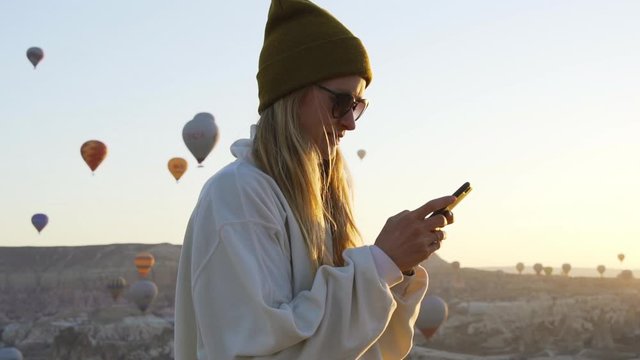 Young blond female traveler blogger texting by smartphone before air balloons